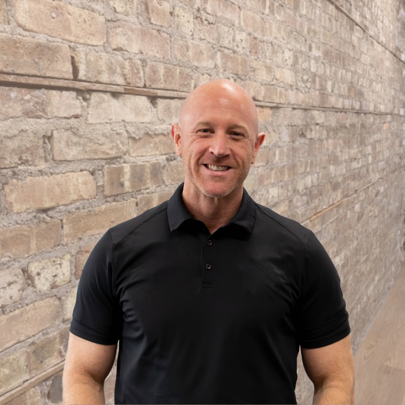 Bald man in black polo smiling against light brick wall at coworking space.