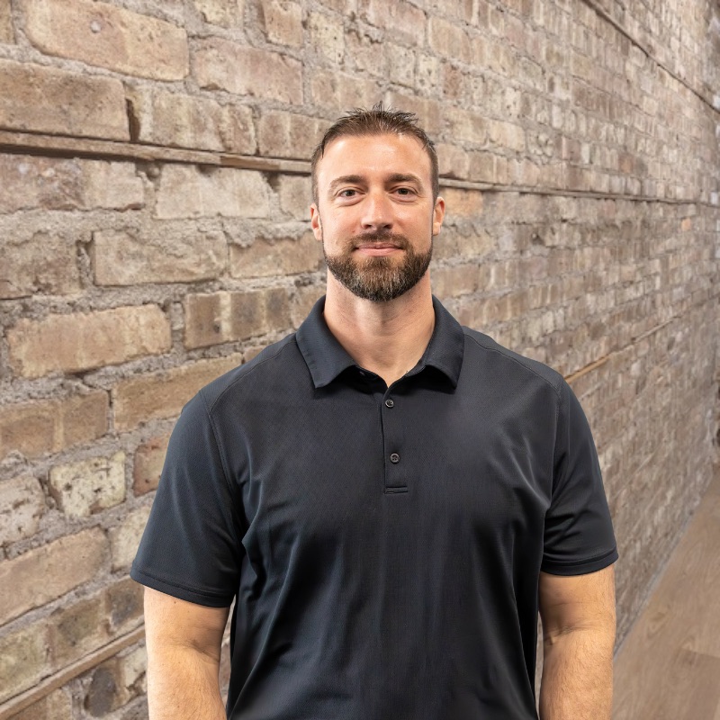 Bearded man with beard in dark polo smiling against light brick wall.