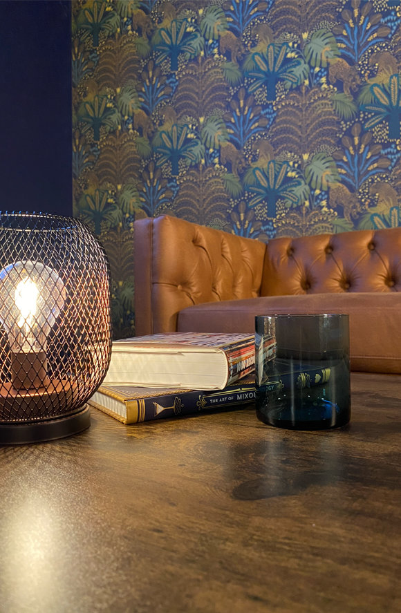 Wooden table with mesh lamp, stacked books, and glass against orange tufted sofa and blue floral wallpaper.