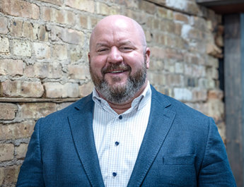 Man with beard in blue blazer and checkered shirt smiling against brick wall at coworking space.