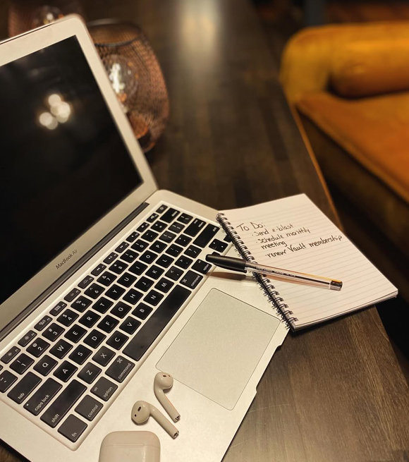 Laptop with to-do list, pen, earbuds, and coffee on wooden table in coworking space.