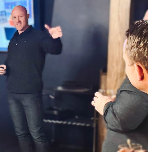Man in black gesturing while presenting to audience member in dark clothing at indoor event.
