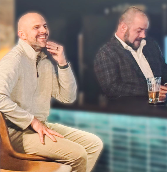 Man in beige quarter-zip smiling while seated, another man in plaid blazer holding drink in background.