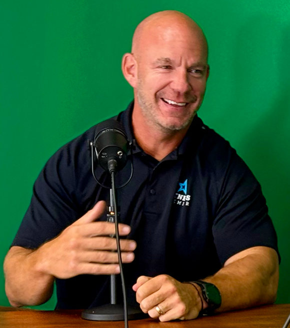 Man gesturing at microphone while speaking, wearing dark polo with logo against green screen background.